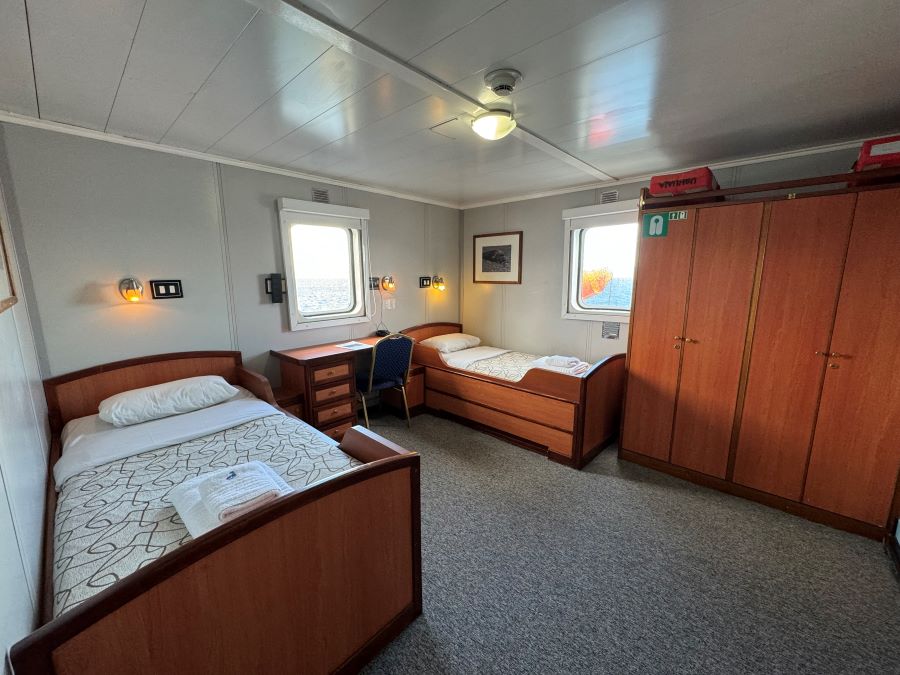 A cozy cabin featuring two single beds with white linens, wooden frames, and gray blankets. The room includes a wooden desk and chair near a window. A spacious wooden wardrobe stands to the right. There is framed artwork on the walls and a ceiling light, perfect for preparing for your Ushuaia Antarctica expedition.