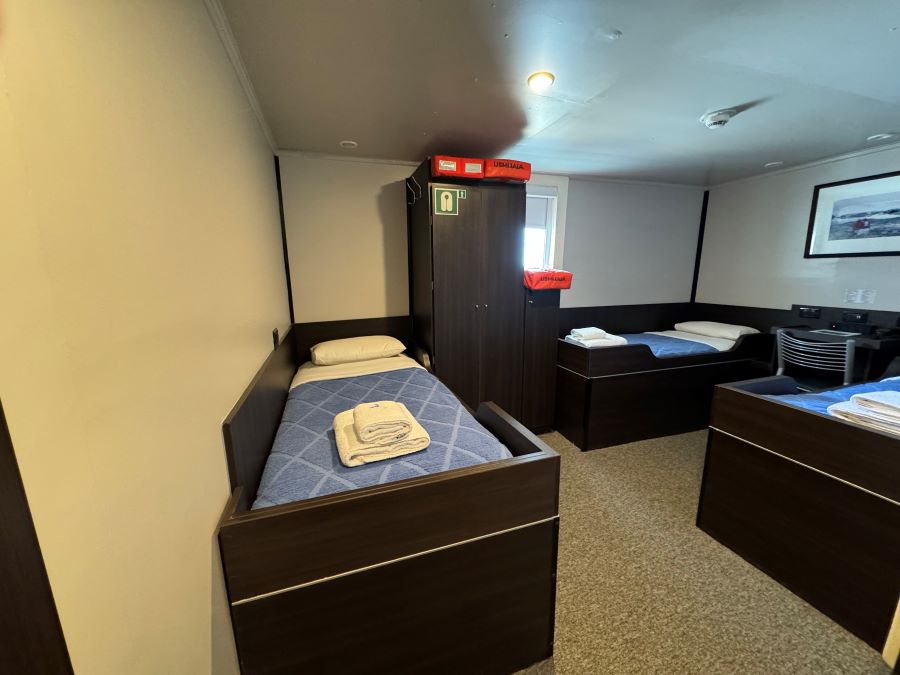 A small room with three single beds made up with blue blankets and white pillows. Towels are neatly placed on the nearest bed. There is a wardrobe in the center and a window in the background, along with a framed picture on the wall. The room feels well-organized and clean, perfect for an Ushuaia Antarctica expedition stay.