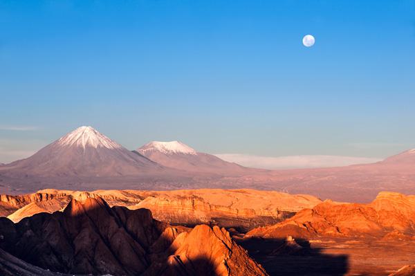 A scenic view of the Atacama Desert at sunset with rugged, red-hued rock formations in the foreground. Snow-capped volcanoes rise in the distance under a clear blue sky, with a full moon visible above the horizon.