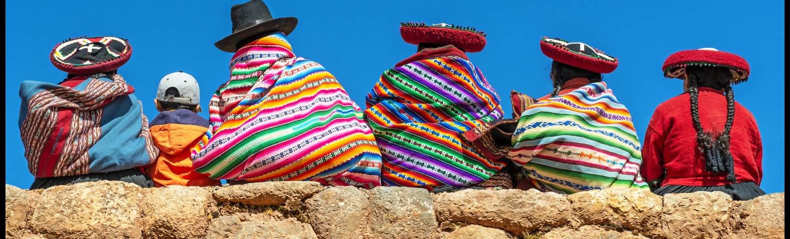 A group of five people wearing traditional, colorful Andean clothing and hats sit on a stone wall. They have their backs to the camera and are set against a bright blue sky.