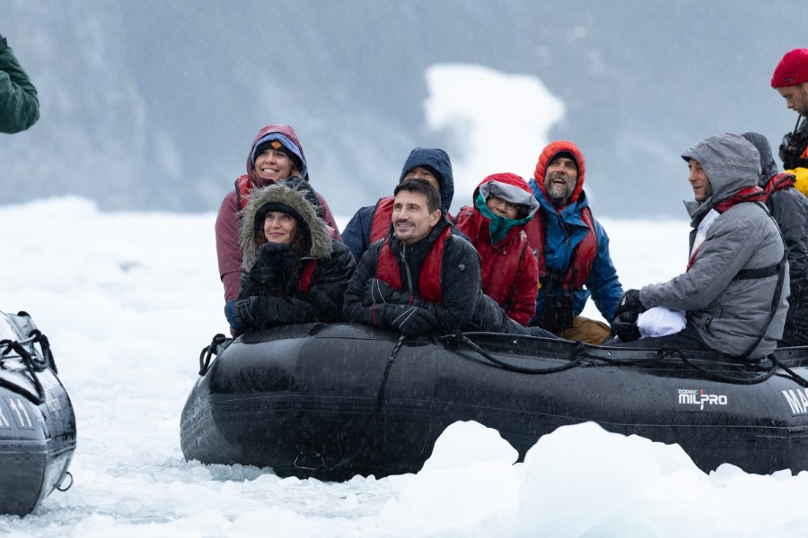 A group of people dressed in winter clothing sits in a black inflatable boat on icy waters. They appear to be enjoying an Antarctica expedition, possibly around a glacier or arctic setting, with snow and ice visible around them.