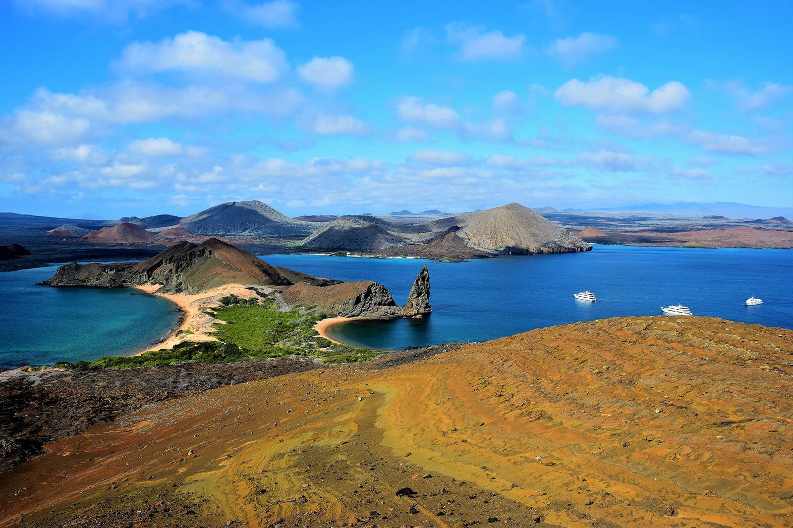 A scenic view of the Galápagos Islands featuring a rugged coastline with blue ocean waters, distinct volcanic formations, and scattered green vegetation. Several boats are anchored near the shore, and the sky is partially cloudy with vibrant blue hues.