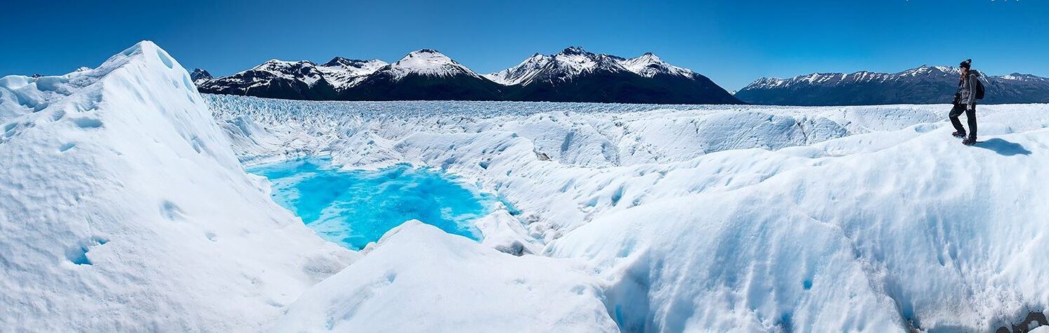 A person stands on a snowy landscape near a bright blue frozen pool on the Perito Moreno Glacier in Argentina, with towering white glaciers and rugged snow-capped mountains in the background under a clear blue sky.