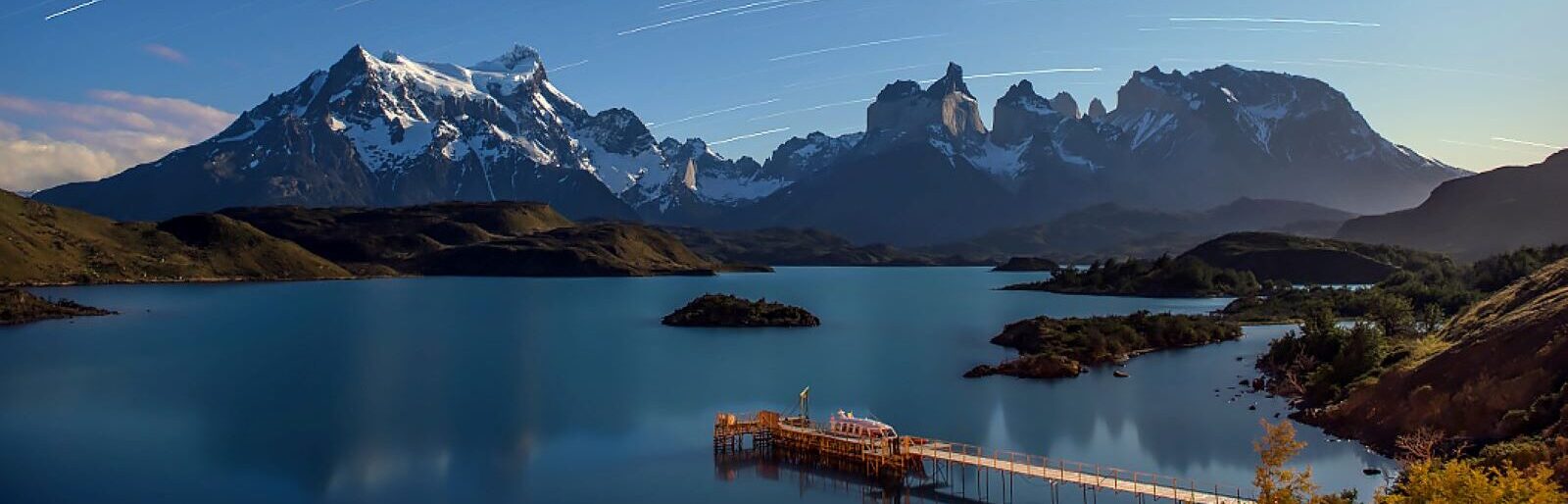 A peaceful lake surrounded by mountainous landscapes under a night sky with star trails. A wooden dock extends into the lake, with a small structure at the end. The distant peaks are partially snow-covered, contrasting with the calm water and clear sky.