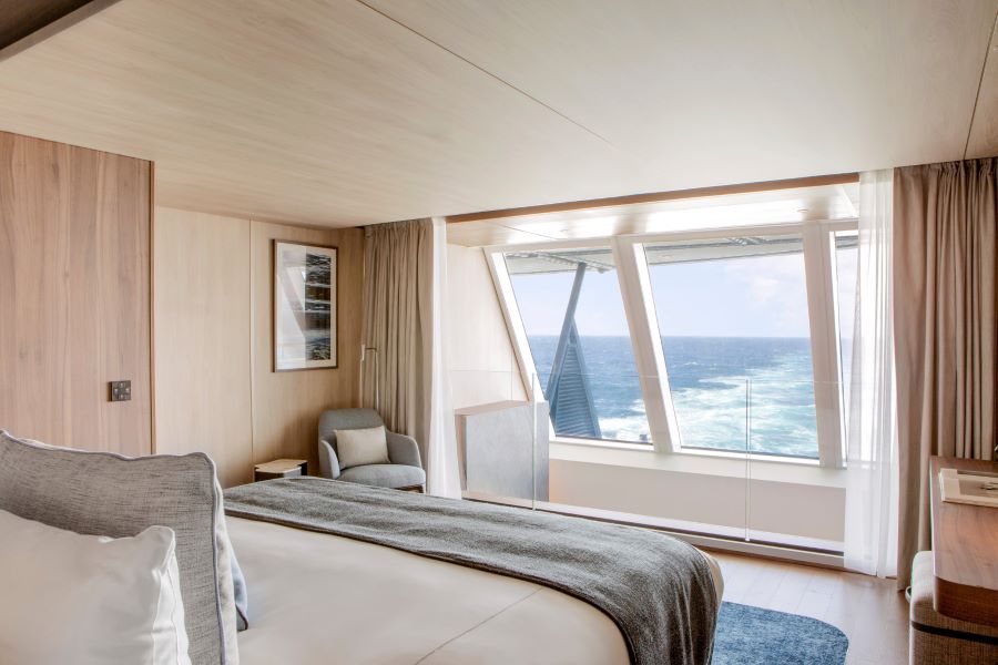 A modern bedroom features a large window offering an ocean view. The room includes a bed with white and gray bedding, a plush armchair, and a framed picture on a light wood-paneled wall. Light curtains frame the window, and the decor is minimalist and serene.