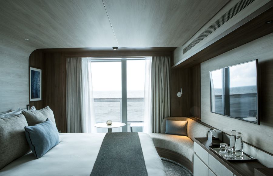 A modern hotel room with a large bed, a gray blanket, and pillows. There is a curved sofa, a wall-mounted TV, a window with curtains and a view of the sea, and a small round table with a slim vase. There is a minimal and serene ambiance with neutral tones.