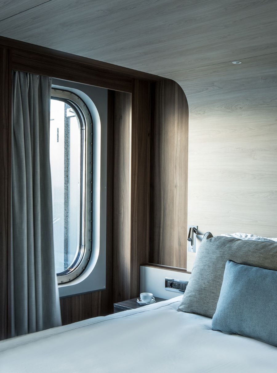 Modern ship cabin interior with a cozy bed and pillows, illuminated by natural light coming through a large porthole window. The wooden accents and soft curtains create a warm and inviting atmosphere. A cup and saucer are placed on the bedside shelf.