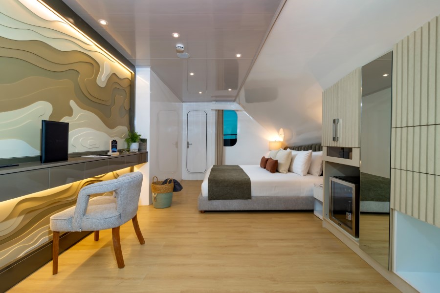 A modern hotel room features a neatly made bed with various pillows, a stylish chair, a desk with a TV, patterned wall art, and a light wood floor. A large mirror, wardrobe, and a window with a view are also present. A woven basket sits beside the bed.