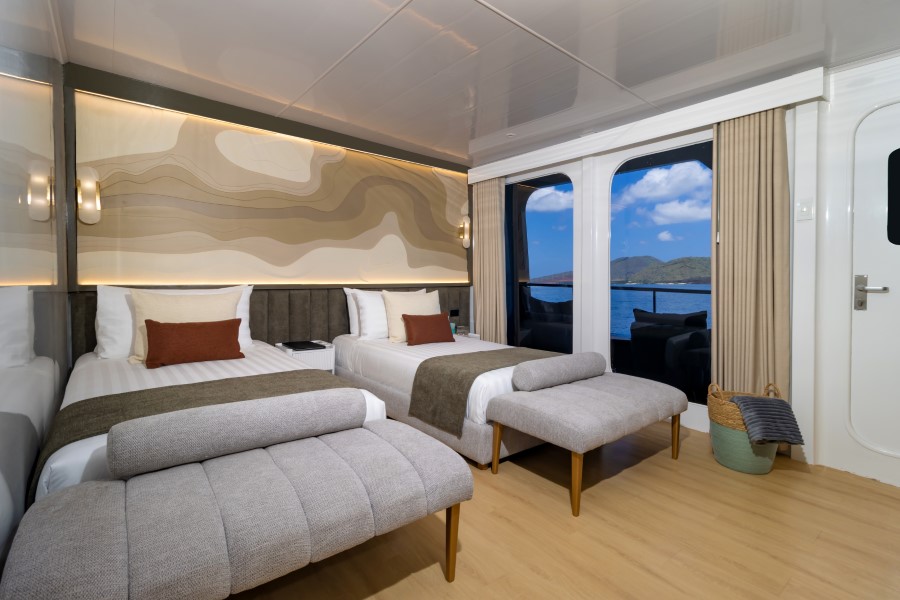 A modern and well-lit cabin featuring two single beds with brown accent pillows, a contemporary patterned wall design, and large windows offering a scenic view of the ocean and mountains. The room has wooden flooring and neutral-toned furnishings, including footstools and wall sconces.