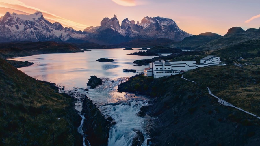 An expansive landscape at sunset features serene waters flowing into a dramatic waterfall. In the foreground, Explora Lodge in Torres del Paine is situated on the edge of the water, surrounded by hilly terrain. Snow-capped mountains tower in the background under a colorful sky.