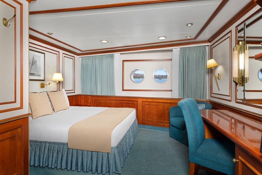 A cozy cabin on the National Geographic Orion expedition ship features a large bed with beige and white bedding, curtain-lined portholes, wooden accents, a blue upholstered chair, and a wooden desk. The room is softly lit with wall-mounted lamps and decorated with simple artwork.