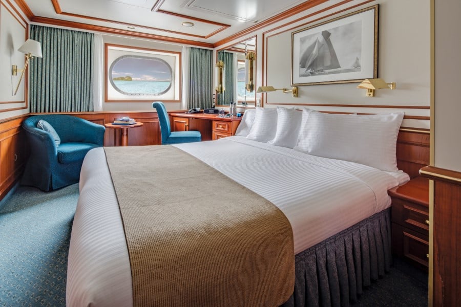 A cozy, well-appointed cruise ship cabin with a neatly made double bed, blue accent chairs, a porthole with a scenic ocean view, a desk, and wooden furnishings. A framed nautical photograph adorns the wall above the bed. Natural light fills the room on this National Geographic Orion expedition ship.