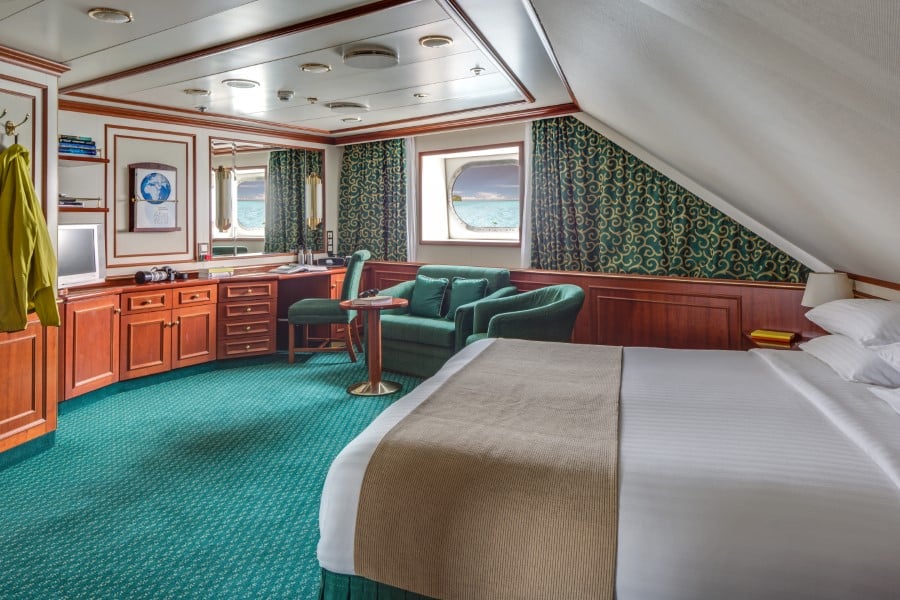 A spacious cruise ship cabin on the National Geographic Orion expedition ship features a large bed, green carpeting, wood-paneled walls, and two windows with ocean views. The room includes a desk, a green armchair with an ottoman, a small table, and wall-mounted storage.