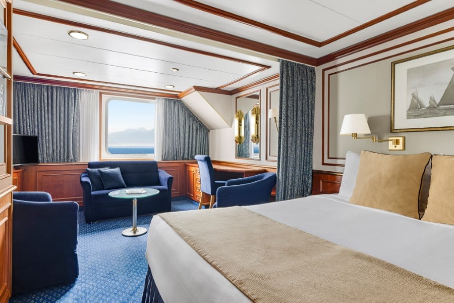 A well-lit, spacious cruise ship cabin on the National Geographic Orion expedition ship features a large bed in the foreground, a seating area with blue armchairs and a sofa, a small glass table, a desk with wooden finishes, and a window revealing a scenic ocean view with distant mountains.