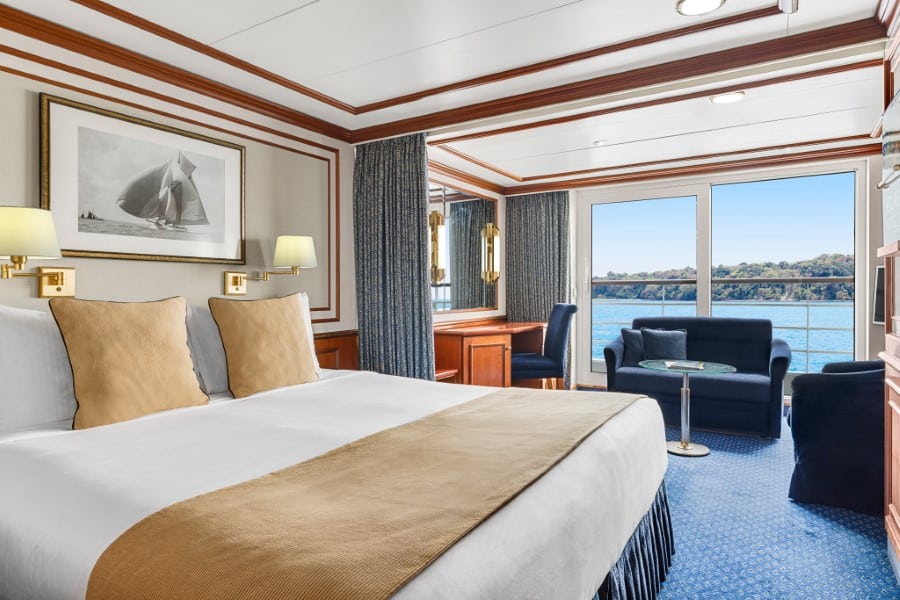 A luxurious National Geographic Orion expedition ship cabin features a large bed with beige pillows and throw, adjacent nightstands with lamps, and a framed sailboat photograph on the wall. There's a seating area with a sofa and table by the window, offering a view of the sea and shoreline.