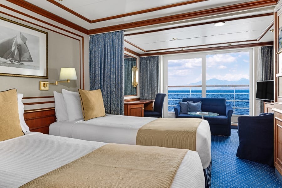 A luxurious cabin on the National Geographic Orion expedition ship features two twin beds, blue and beige decor, a large window with an ocean view, and a seating area with a small table and two chairs. The cabin is well-lit with a framed sailboat photo on the wall and curtains drawn to the side.